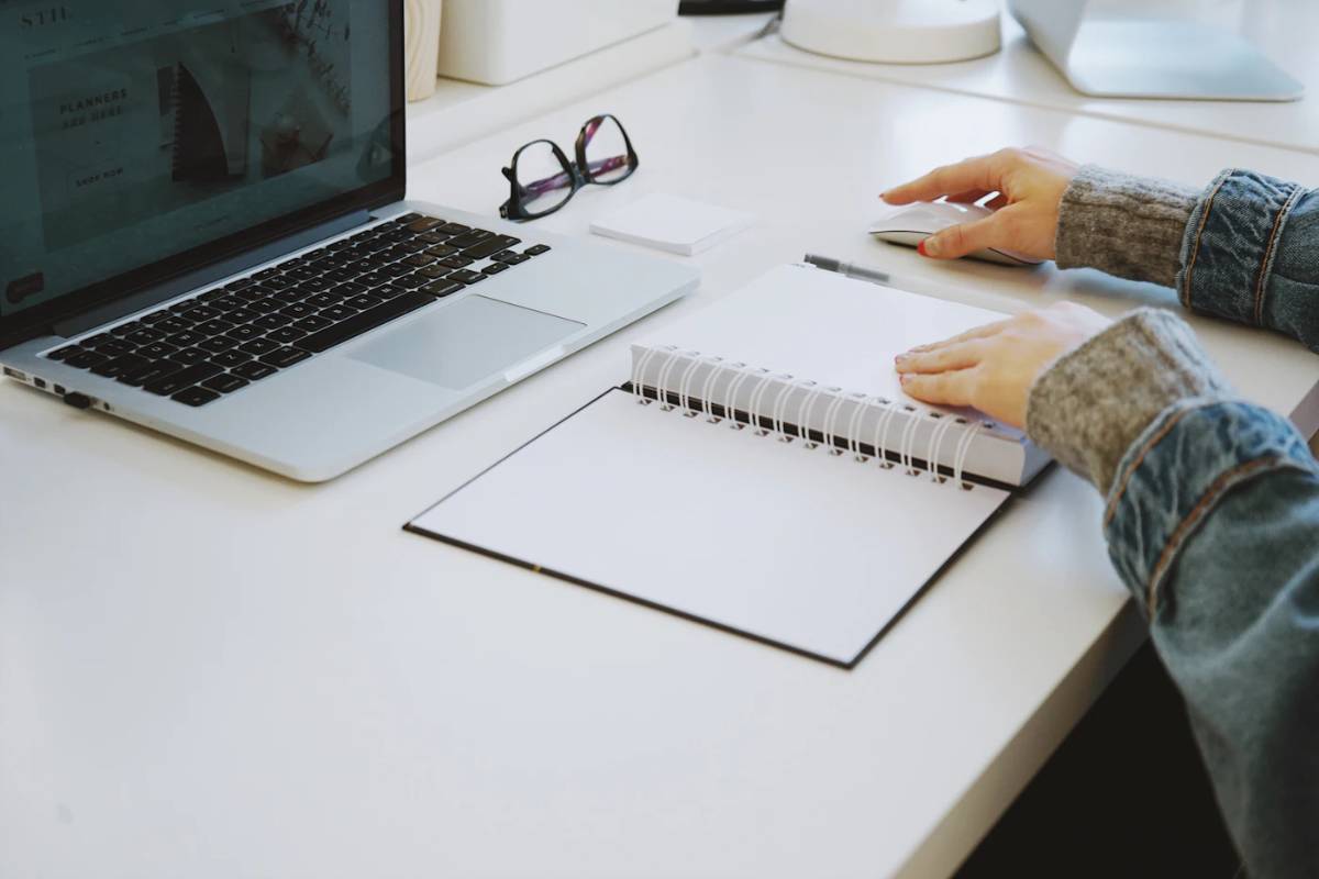 Hands at a white desk with a laptop and an open notebook